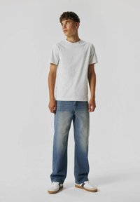 Camiseta de algodón gris claro combinada con jeans rectos de un azul medio. El modelo lleva zapatillas blancas con rayas negras. Diseño simple y casual.