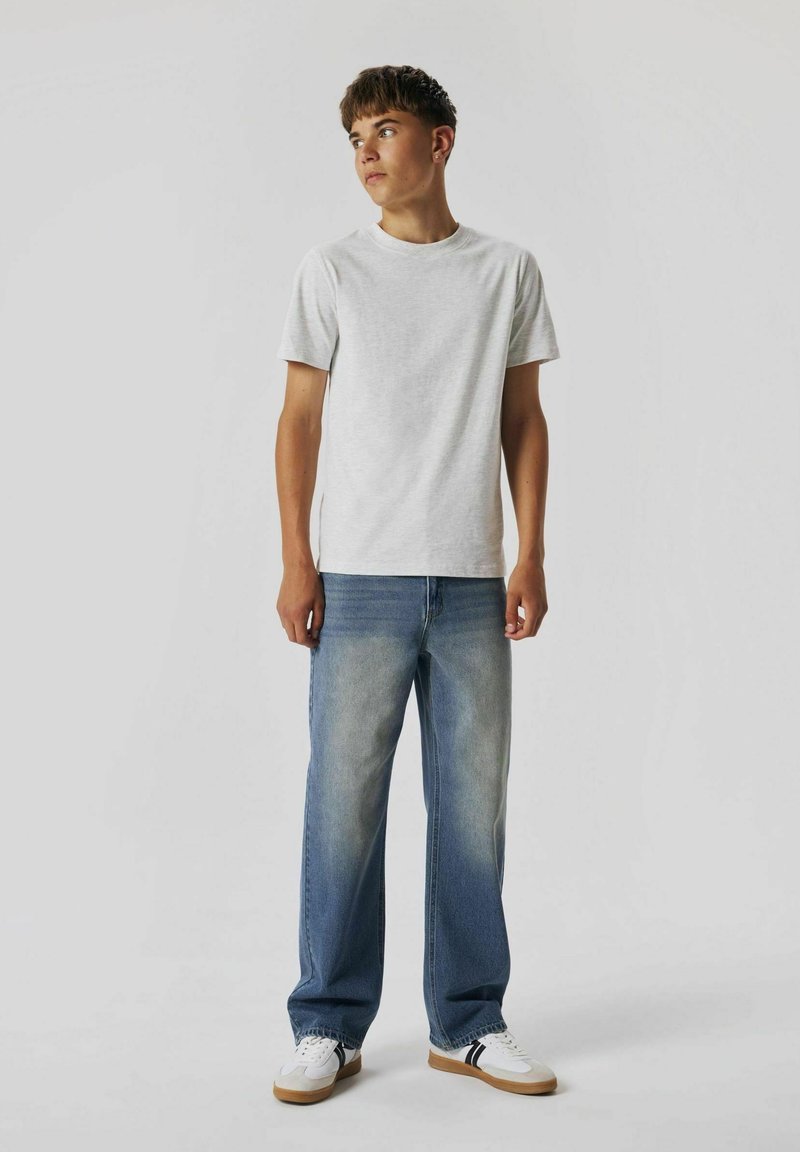 Camiseta de algodón gris claro combinada con jeans rectos de un azul medio. El modelo lleva zapatillas blancas con rayas negras. Diseño simple y casual.