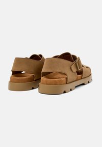 Brown suede sandals with a flat, thick sole. Features a wide adjustable strap and an open back design, showcasing a soft interior.