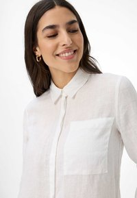 White linen shirt with a button-down collar, front pocket, and a lightweight, textured fabric. Model with long, dark hair and hoop earrings.