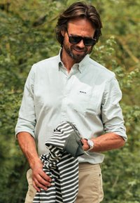 Lichtgroen overhem shirt met lange mouwen, lichte stof en een zak; houdt een opgevouwen zwart-wit gestreept trui vast; buitensetting.