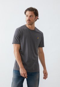 Tricou din bumbac gri, cu decolteu rotund, având un mic logo galben brodat pe piept. Asortat cu blugi albastri decolorati.
