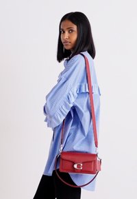 Červená kožená crossbody taška so strieborným uzáverom, obdĺžnikový tvar, hladká textúra a nastaviteľný popruh; nosená cez modrú prúžkovanú košeľu.