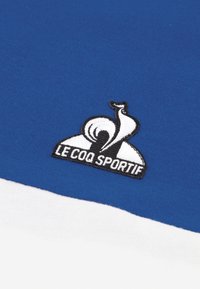 Tejido de algodón azul con una raya blanca, con un logo bordado en blanco y negro de Le Coq Sportif. La textura es suave y ligera.