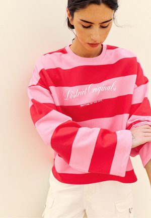 Junge Frau trägt einen pink-rot gestreiften Pullover mit dem Text "District Originals" und richtet den Ärmel vor einem einfarbigen beigen Hintergrund.