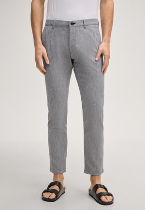 Mężczyzna ubrany w przylegające szare spodnie, białą koszulkę włożoną do środka i czarne sandały na podwójne paski, stojący na neutralnym tle.