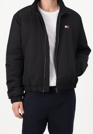 Mand iført en sort Tommy Jeans lynlåstrøje over en hvid t-shirt med den ene hånd i lommen mod en ensfarvet baggrund.