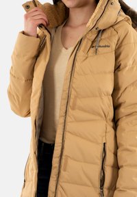 Giacca imbottita beige con colletto foderato in pelliccia, chiusura con zip e tasche laterali. Tessuto testurizzato con una finitura liscia e logo Columbia sul petto.