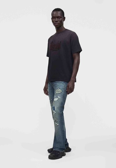 Άνδρας που φοράει μαύρο T-shirt με σχέδια, φθαρμένο μπλε τζιν με επιδιορθώσεις και μαύρα αθλητικά, στέκεται μπροστά σε ένα ομοιόχρωμο φωτεινό φόντο.