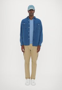 Denim shirt in blue with two chest pockets, layered over a light blue T-shirt. Tan cargo pants and white sneakers complete the outfit.