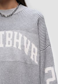 Szary sweter z dzianiny z teksturowanym wzorem, z białym napisem i akcentami. Żebrowany dekolt i różne wzory materiałów.