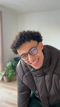Een persoon met krullend haar draagt ronde, bruine montuurbrillen en een zwarte pufferjas met een ritssluiting, leunend naar voren tegen een lichte achtergrond.