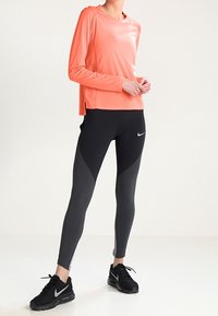 Langärmliges, leuchtend korallenrotes Sportoberteil mit Seitenschlitzen, kombiniert mit schwarzen Leggings mit grauen Akzenten und einem reflektierenden Nike-Logo.
