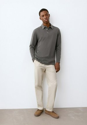 Joven de pie contra una pared blanca, vistiendo un polo gris de manga larga, pantalones beige y zapatos casuales marrones, con una mano en el bolsillo.