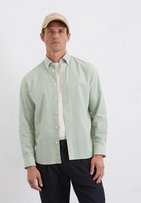 Hellgrünes gestreiftes Hemd mit Button-Down-Kragen, langen Ärmeln und weißen Knöpfen, über einem cremefarbenen Rundhals-Shirt getragen. Schwarze Hose.