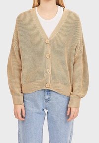 Beżowy sweter z dzianiny z dekoltem w kształcie litery V, pięcioma drewnianymi guzikami i luźnym krojem. Posiada ściągacze u dołu i szerokie rękawy, zestawiony z jasnoniebieskimi dżinsami.