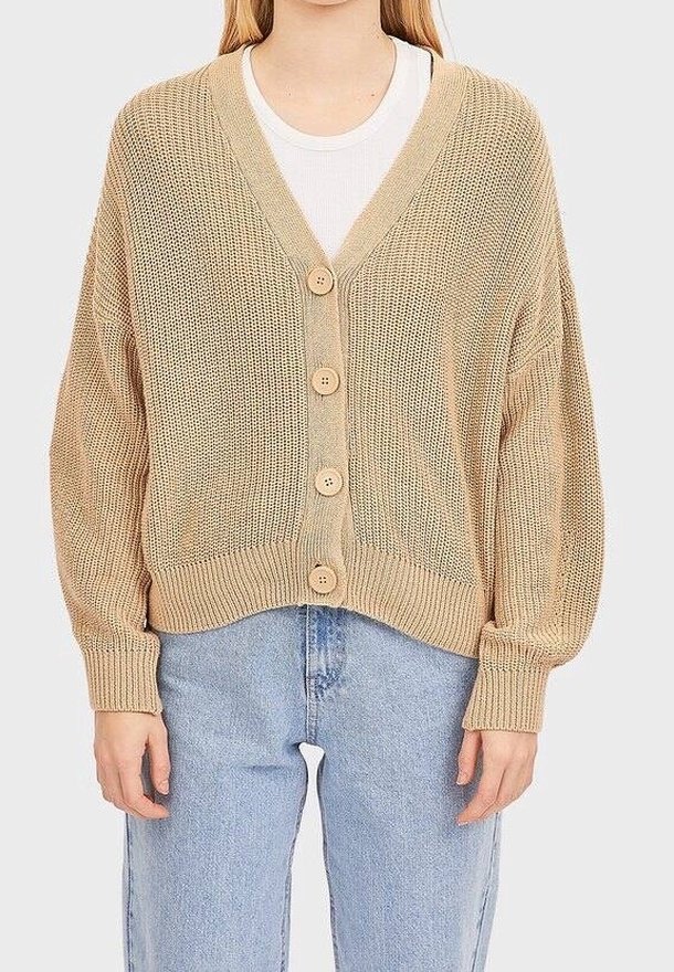 Beżowy sweter z dzianiny z dekoltem w kształcie litery V, pięcioma drewnianymi guzikami i luźnym krojem. Posiada ściągacze u dołu i szerokie rękawy, zestawiony z jasnoniebieskimi dżinsami.