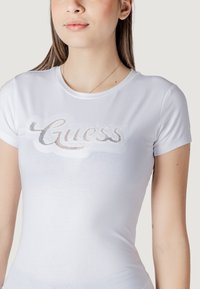 Witte katoenen t-shirt met korte mouwen met een strass "Guess" logo in cursief op de borst. Glad textuur, getailleerd ontwerp.