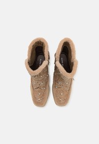 Bottines en daim beige avec un col en fausse fourrure doux, ornées de clous argentés et d'accents turquoise. Semelles profil bas et fermeture à lacets.