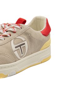 Sneaker in suede beige con dettagli rossi, lacci bianchi a trama, suola gialla e un design del logo sul lato. Forma arrotondata con collarino imbottito.