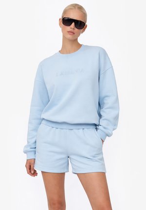 Kobieta ubrana w jasnoniebieski zestaw z bluzy i spodenek, pozująca z jedną ręką w kieszeni i oversize’owymi czarnymi okularami przeciwsłonecznymi.