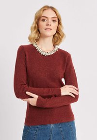 Burgundijas krāsas džemperis ar teksturētu apdari, kuru rotā dārgakmeņu apkakle. Garas piedurknes ar pieguļošiem aprocēm, kombinēts ar zilu džinsus svārkiem.