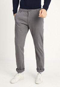 Uomo vestito con pantaloni grigi arrotolati, sneaker bianche e una maglietta a maniche lunghe blu navy, in piedi con le mani nelle tasche.