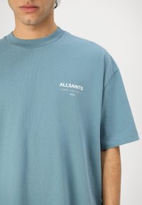 T-shirt en coton bleu clair avec des manches courtes. Présente un logo « ALLSAINTS » blanc sur la poitrine. Design simple, texture douce, coupe standard.