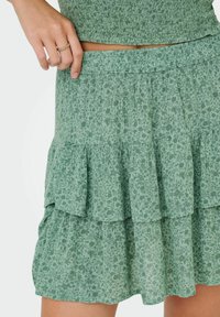 Mujer vestida con una falda de flores verde en capas y un top a juego, con la mano descansando en la cinturilla y un anillo de oro en el dedo.