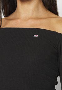 Schwarzes, geripptes Off-Shoulder-Oberteil mit kleinem gesticktem Logo auf der linken Seite. Glatte Textur mit tailliertem Design.