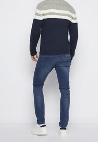 Strickpullover mit einem Farbblockdesign in Grau, Weiß und Marineblau. Getragen mit dunkelblauen Jeans und weißen Turnschuhen. Strukturierter Stoff mit gestreiften Bündchen.