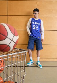 Maglia da basket blu con la scritta "FRANCE" e il numero 32, pantaloni corti in denim, camicia a maniche lunghe bianca e sneaker multicolori. Palloni da basket in un carrello di metallo.