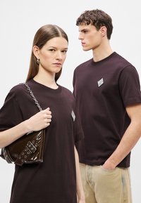 Dwaj modele noszą ciemnobrązowe koszulki z białym paskiem na lewej piersi. Jeden z modeli trzyma brązową lakierowaną torebkę na łańcuszkowym pasku.