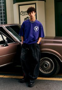 Camiseta oversized morada con un logo, jeans sueltos negros y zapatos de plataforma negros, de pie junto a un coche marrón con ruedas plateadas.