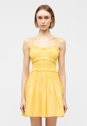 Mujer joven con un vestido amarillo sin mangas y fruncido, con detalles de volantes, de pie frente a un fondo liso de color gris claro.