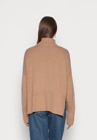 Beżowy oversize'owy sweter dzianinowy z wysoką szyją, ściągaczem u dołu oraz bocznymi rozcięciami, w połączeniu z niebieskimi dżinsami o prostym kroju.