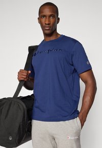 Navyfarbener Baumwoll-T-Shirt mit gesticktem "Champion"-Schriftzug. Das Modell hält eine schwarze Sporttasche. Graue Jogginghose mit Logo-Akzent. Schlichtes Design.