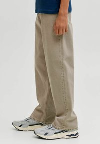 Jeans de corte ancho beige con un acabado texturizado, que cuentan con un bolsillo lateral, combinados con zapatillas deportivas grises y marino. El diseño es de corte relajado.
