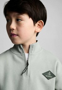 Lichtgroene ritsvest met hoge kraag, voorzien van een voorkantzak en een donkergroen logo-patch in de vorm van een diamant.