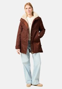 Ruskea eristetty parkatakki, jossa on tekoturkista vuorattu kaulus, vetoketju- ja napsulukot, etutaskut ja hyvin istuva leikkaus. Pukeutuneena vaalean kermanvärisen paidan päälle.