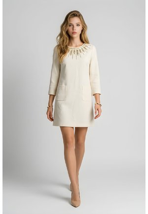 Mujer rubia con vestido color crema con escote adornado, mangas 3/4, bolsillos delanteros, pulseras doradas y zapatos de tacón en tonos nude.