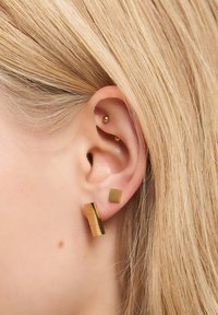 Nahaufnahme eines blonden Ohres mit vier goldenen Ohrringen: einem kleinen quadratischen Stecker, einem gestapelten rechteckigen Pendant und zwei kleinen goldenen Kugel-Piercings.