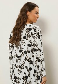 Langærmet bluse i let hvidt stof med et sort blomstermønster. Nøglefunktioner inkluderer knaplukning ved håndleddene og en afslappet pasform.