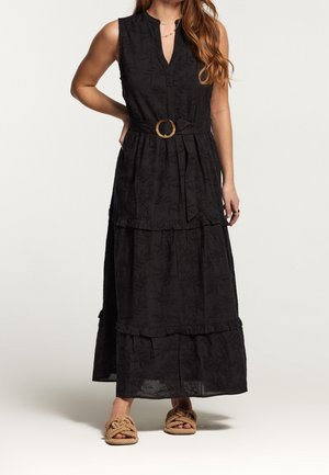 Zwarte mouwloze maxi-jurk met riem en gestructureerd bloemmotief, gedragen met beige geweven open-toe sandalen, model met hand op de heup.