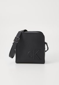Black leather crossbody bag with embossed "CK" logo. Features a zip closure, adjustable strap, and angular shape. Smooth texture.