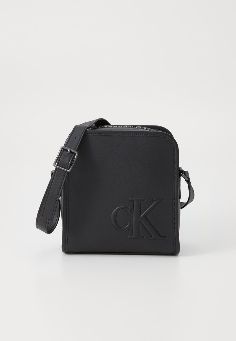 Black leather crossbody bag with embossed "CK" logo. Features a zip closure, adjustable strap, and angular shape. Smooth texture.
