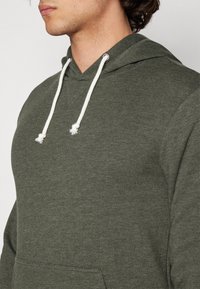 Sudadera de color verde oliva hecha de una tela suave, con un bolsillo frontal, cordones blancos y un diseño de cuello en V.