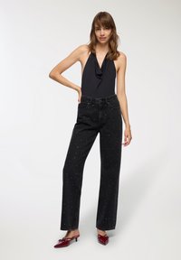 Czarny bezrękawnik z dekoltem w stylu drapowanym, zestawiony z czarnymi dżinsami z wysokim stanem, ozdobionymi białymi akcentami rozmieszczonymi losowo oraz prostym krojem.