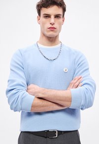 Világoskék bordázott kötött pulóver hosszú ujjakkal, kerek nyakkal, ezüst lánc nyaklánccal és egy kör alakú logóval a mellkason.