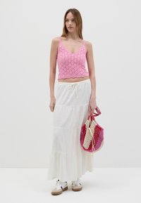 Top crop all'uncinetto rosa, gonna maxi bianca a più strati e borsa intrecciata multicolore. Modella che indossa sneakers con suola color sabbia.